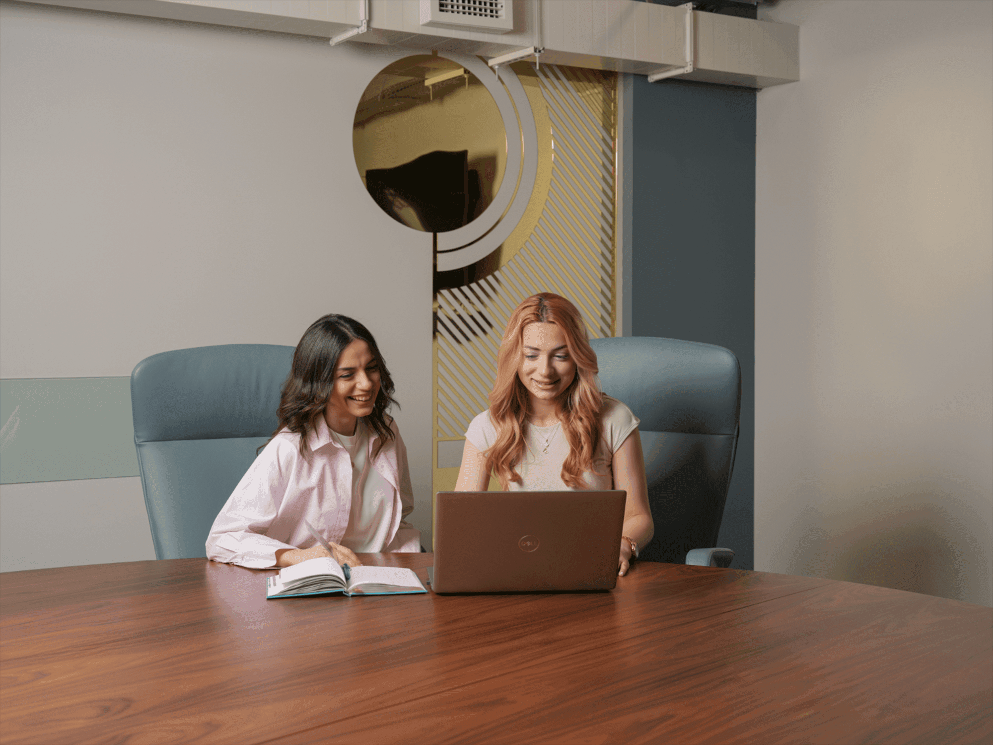 Two smiling women collaborating at a conference table with a laptop and an open book.