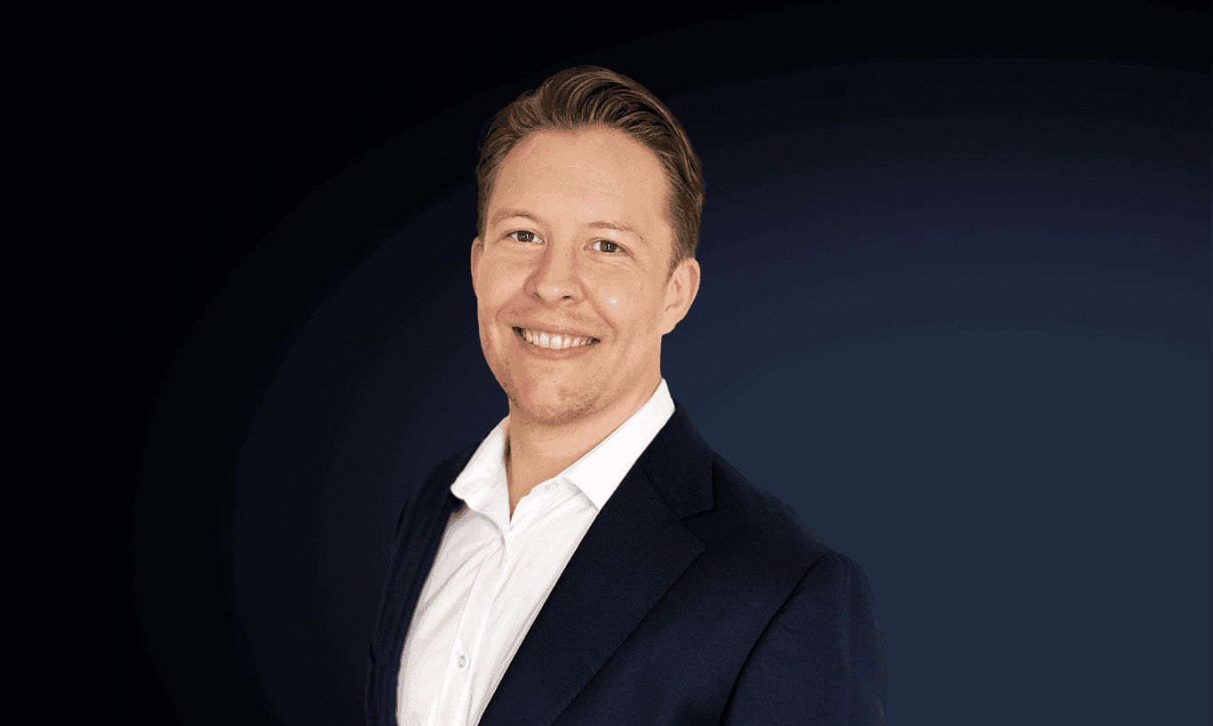Smiling man in white shirt and dark suit jacket against a dark background.